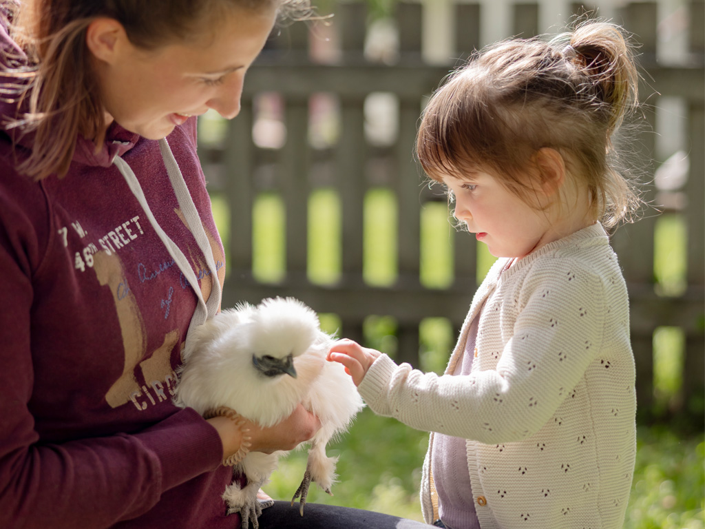 Mutter hält weisses Huhn auf dem Arm und Kind streichelt das Huhn